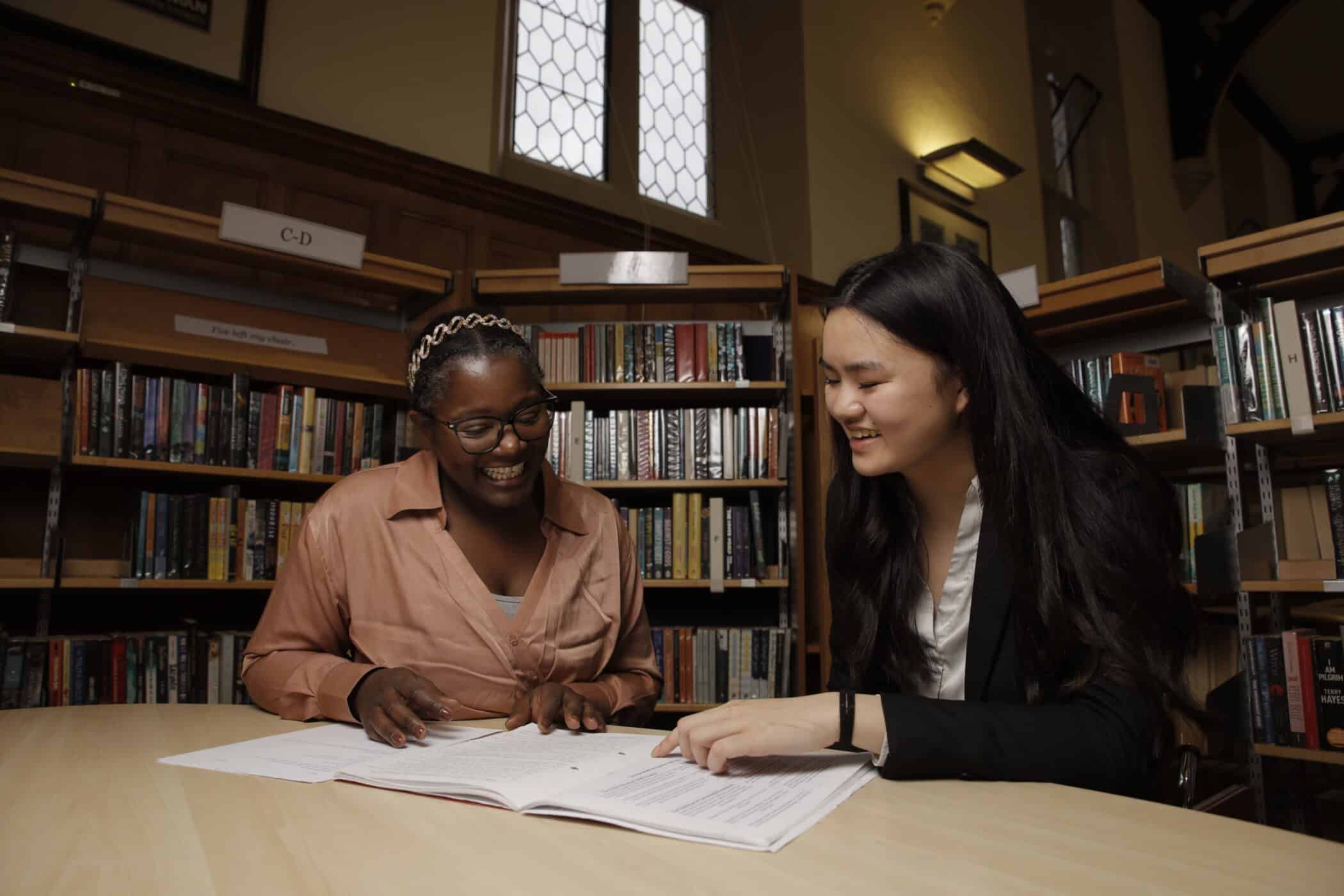 Two students sit at a round table in a library, smiling and discussing open documents in front of them. Bookshelves filled with books surround them, and soft natural light enters through tall, patterned windows in the background.