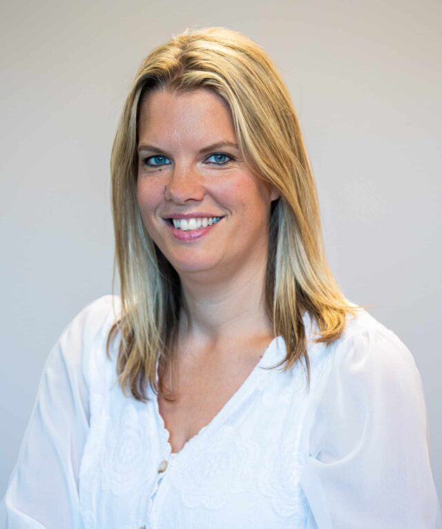 Smiling woman with shoulder-length blonde hair wearing a white blouse, facing the camera in a professional head-and-shoulders portrait against a softly blurred neutral background.