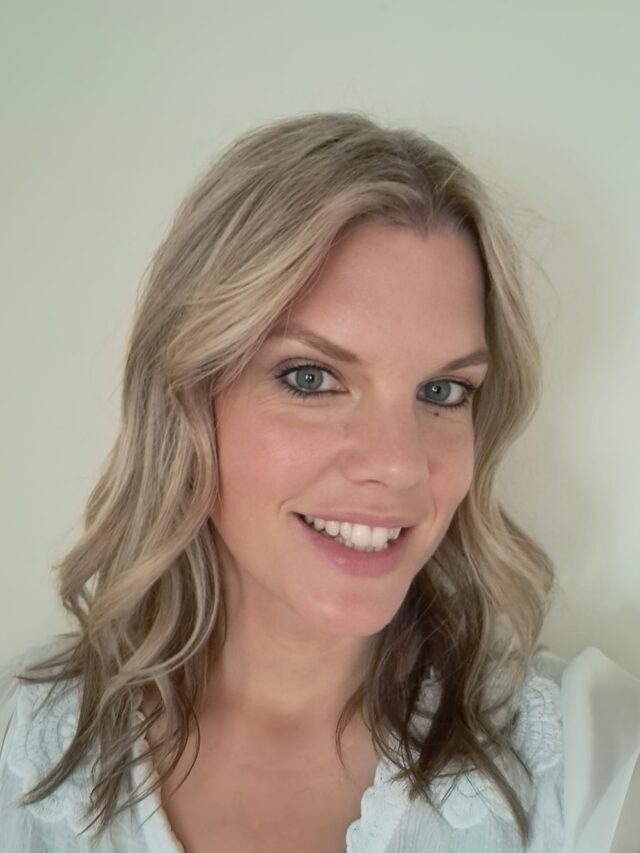 Smiling woman with shoulder-length ash blonde hair styled in loose waves, wearing a white blouse with a scalloped collar. Natural makeup highlights blue-grey eyes. Head-and-shoulders portrait against a plain light background.