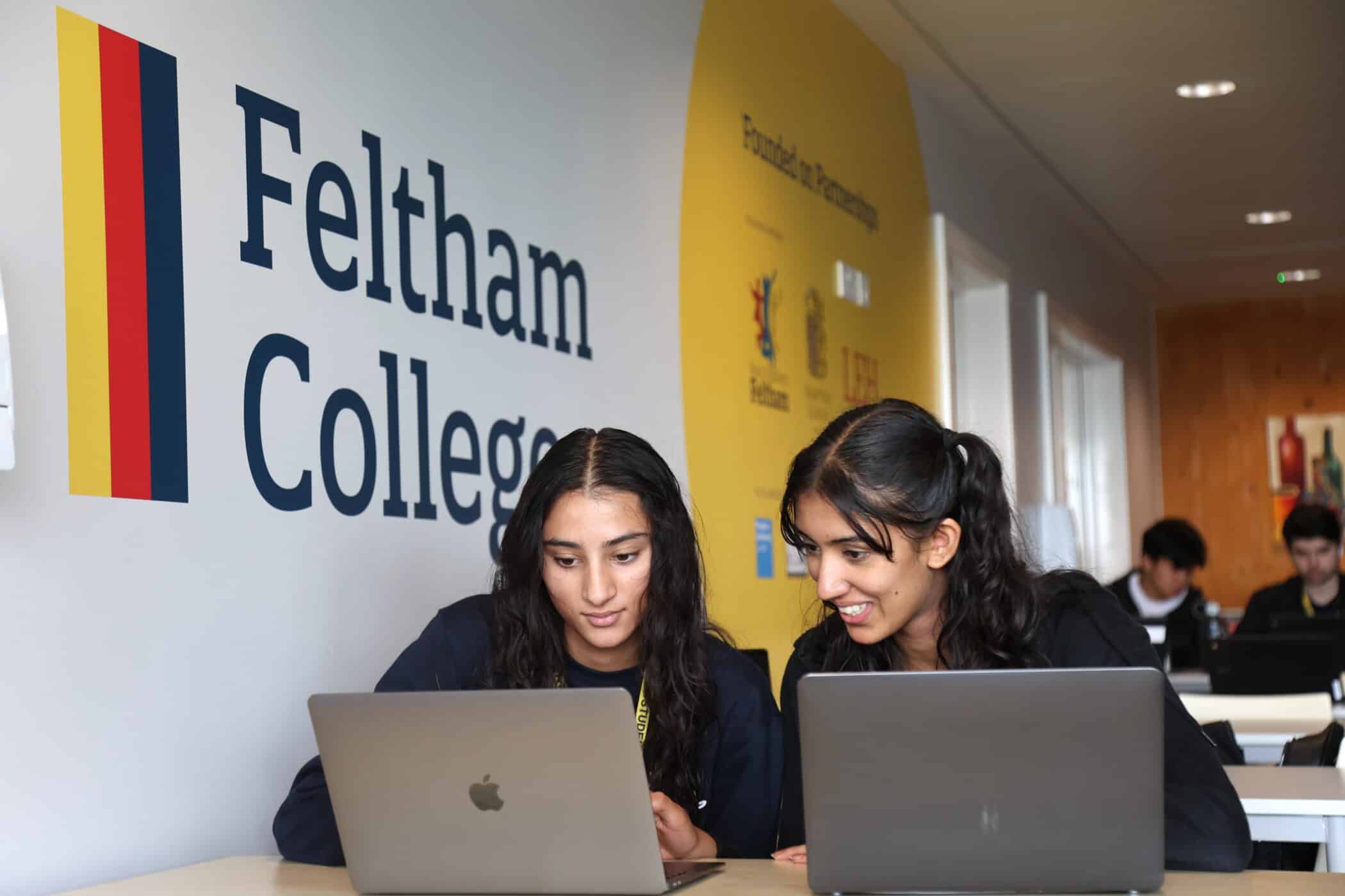 Two female students sit side by side at a desk, working on laptops in a modern study area with a large