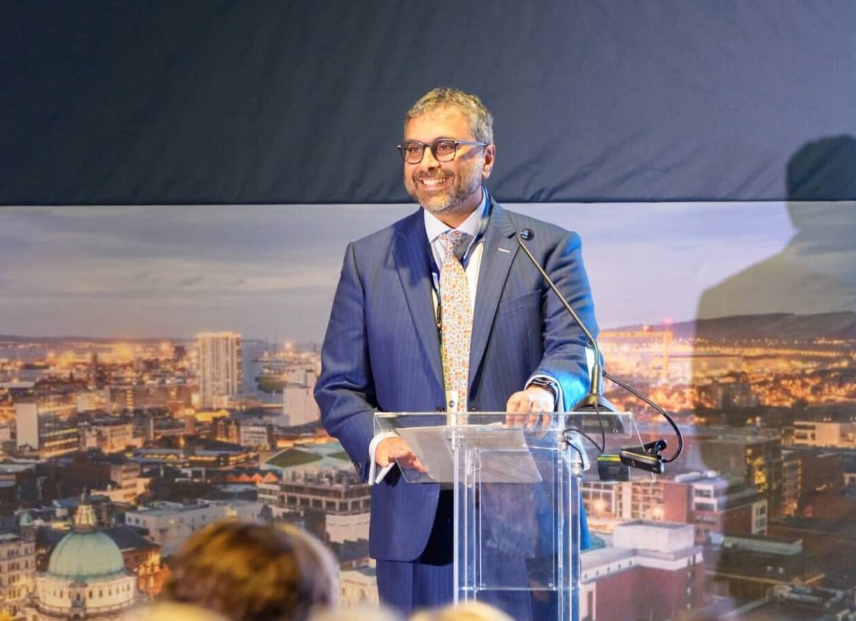 A man in a blue suit, patterned tie, and glasses stands at a clear podium, delivering a speech with a smile. He is positioned in front of a large backdrop featuring a cityscape with illuminated buildings at dusk. A microphone is attached to the podium, and an audience is visible in the foreground. His shadow is cast on the backdrop.