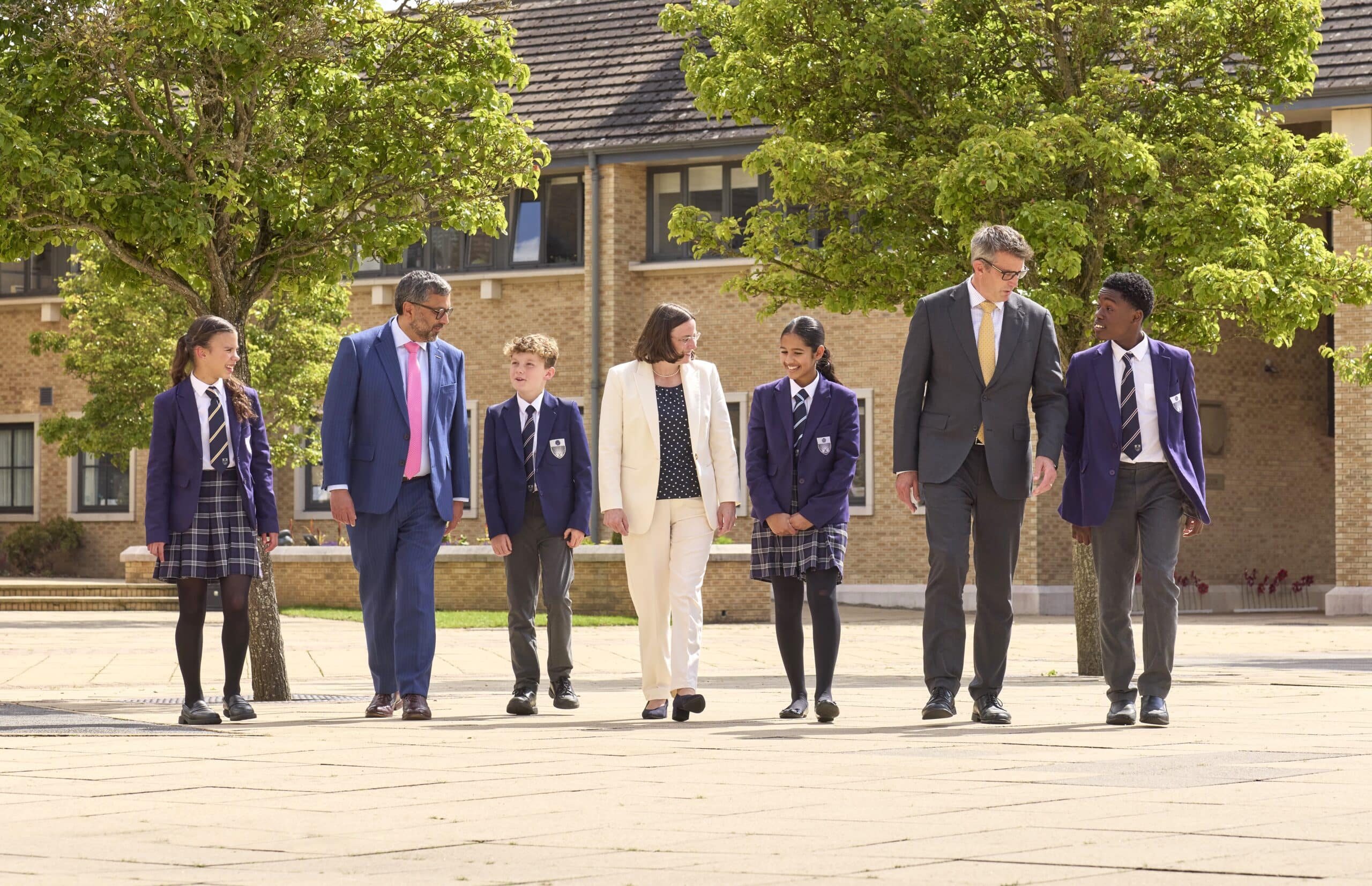 Group of students in purple blazers walking outdoors with three teachers dressed in professional attire, engaged in friendly conversation on a school campus surrounded by trees and modern brick buildings.