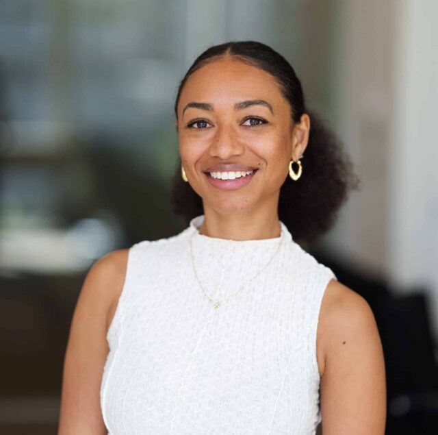 Woman smiling in an office setting, wearing a sleeveless white textured top, gold hoop earrings, and a delicate necklace with a small charm. Her hair is styled in a puff and pulled back from her face. The background is softly blurred.