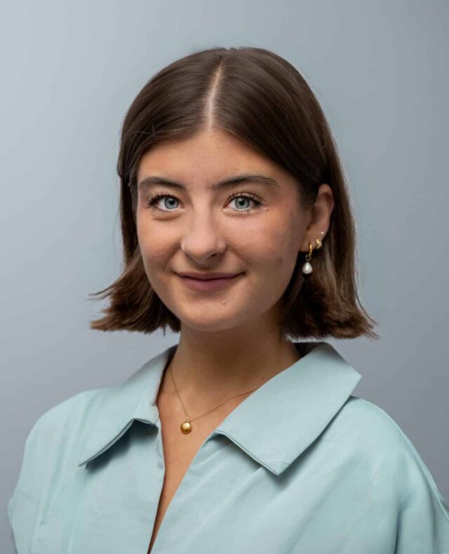 A woman with a short, dark brown bob and blue eyes, wearing a pale blue collared blouse, gold necklace and pearl drop earrings. She smiles gently at the camera against a plain grey background.