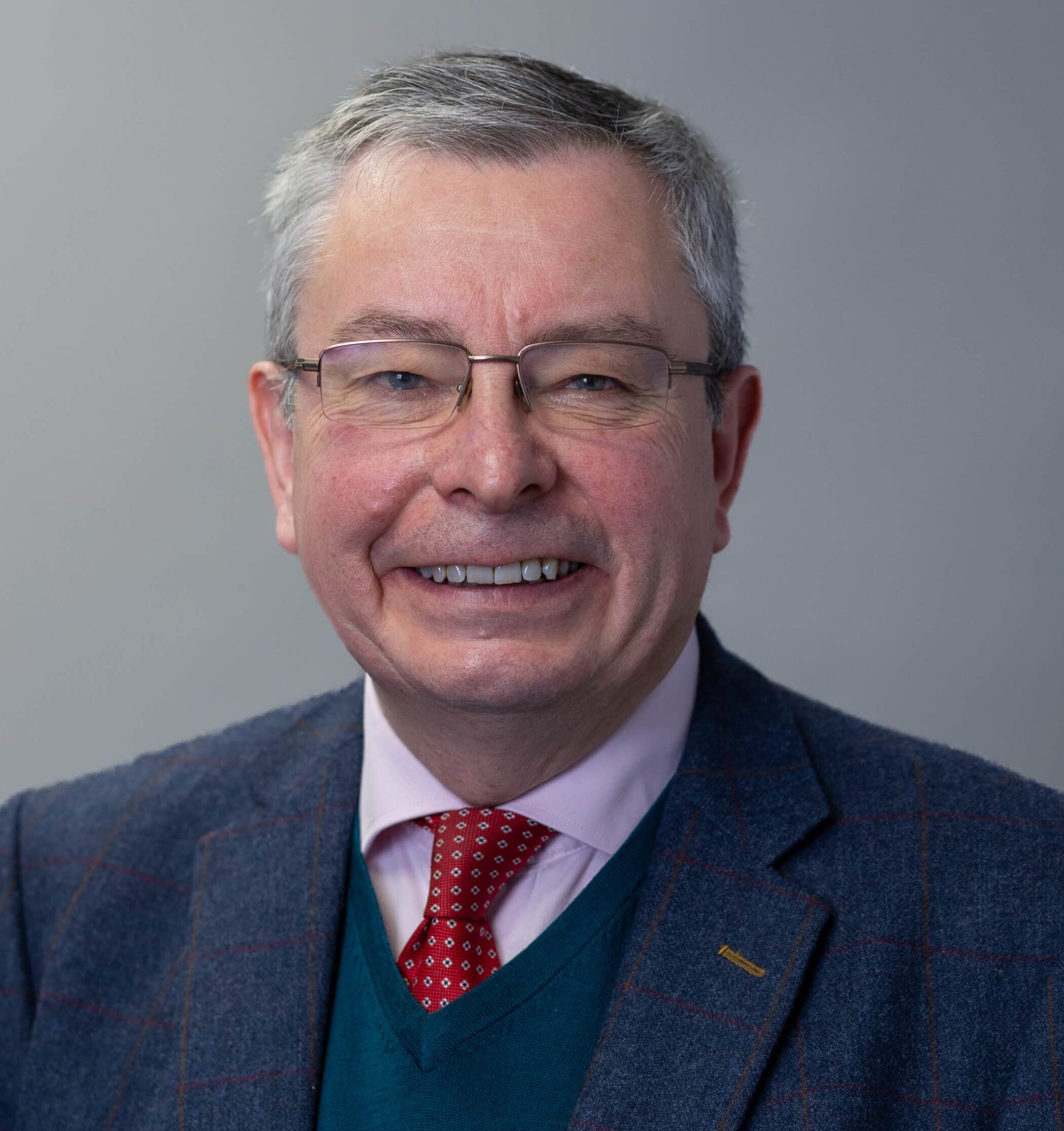 Smiling man with short grey hair and glasses, wearing a checked suit jacket, pale shirt, blue jumper and red patterned tie, posed against a plain grey background.