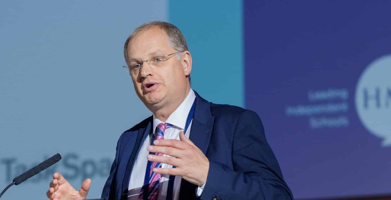 Man in a suit and glasses speaking on stage, gesturing with one hand. A presentation slide with a blue background is visible behind him in a conference setting.