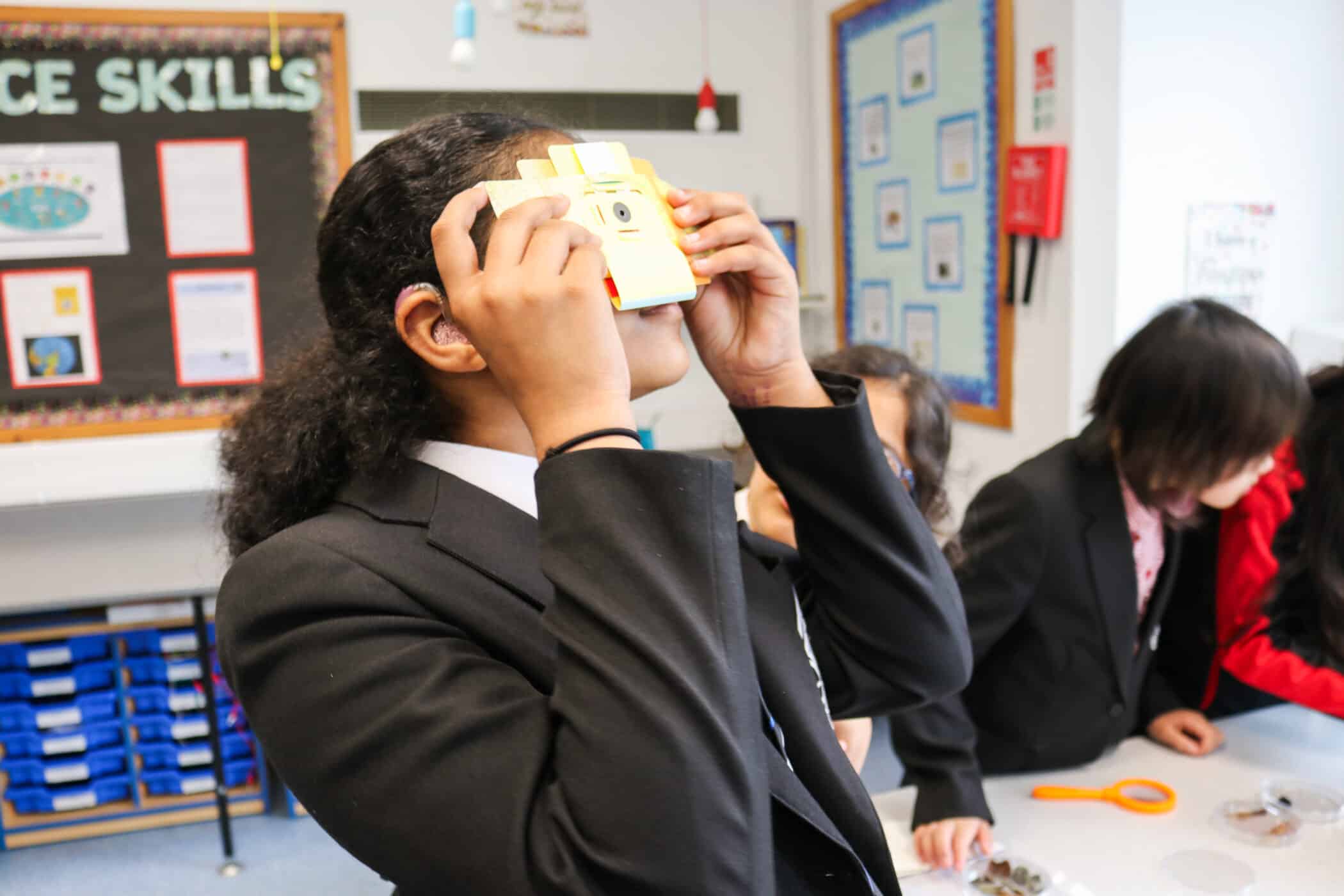 A student wearing a black school blazer and a white shirt is using a small yellow virtual reality (VR) viewer in a classroom. She has dark, curly hair and is wearing a hearing aid in her left ear. Other students in black blazers are engaged in activities in the background. The classroom walls are decorated with educational posters, and a bulletin board with 