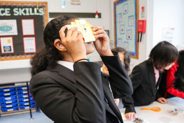 A student wearing a black school blazer and a white shirt is using a small yellow virtual reality (VR) viewer in a classroom. She has dark, curly hair and is wearing a hearing aid in her left ear. Other students in black blazers are engaged in activities in the background. The classroom walls are decorated with educational posters, and a bulletin board with 