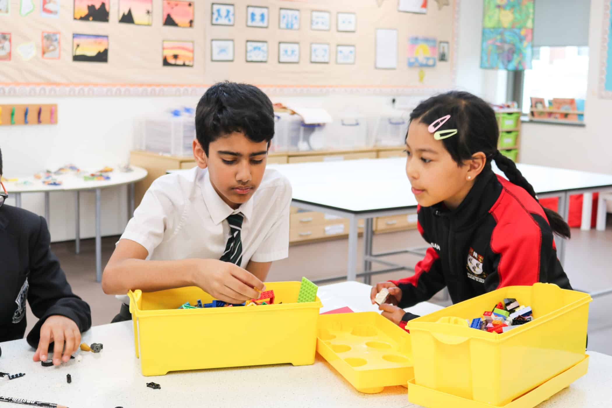 A group of students in a classroom are engaging in a hands-on learning activity using colorful building blocks. A boy in a white shirt and striped tie is focused on selecting pieces from a yellow container, while a girl in a red and black sports jacket with hair clips watches attentively. The classroom background features artwork and educational materials on the walls, with additional storage containers and tables in the space.