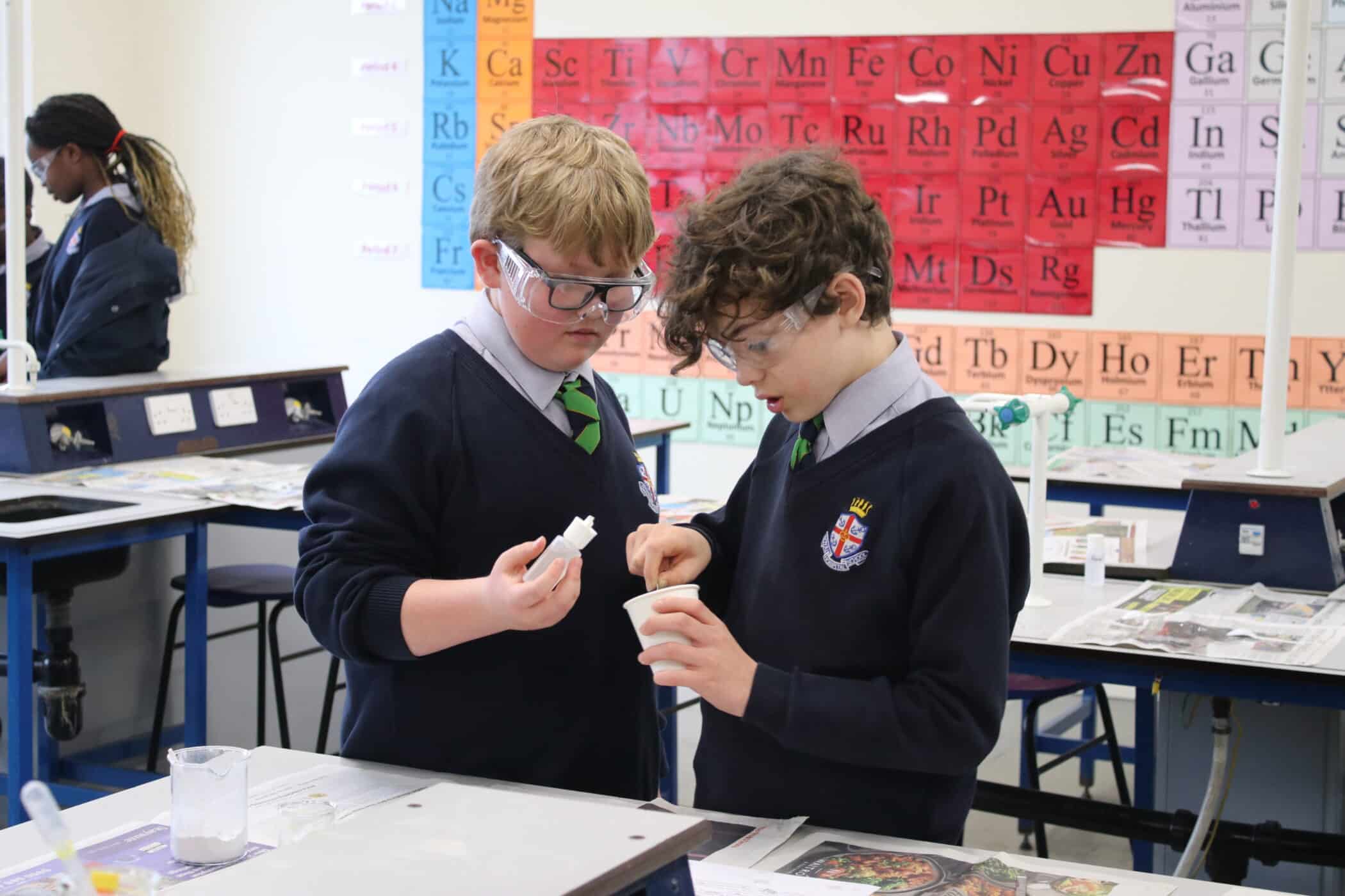 wo male students in navy blue school uniforms, striped green ties, and safety goggles work together on a hands-on science experiment in a classroom. One pours a substance into a cup while the other observes. Behind them, a colorful periodic table is displayed on the wall. A speech bubble overlay reads: 