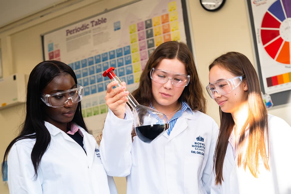 Three female students in white lab coats and safety goggles conduct a chemistry experiment in a school science lab. One student holds a flask containing a dark liquid while the others observe. A speech bubble overlay reads: 