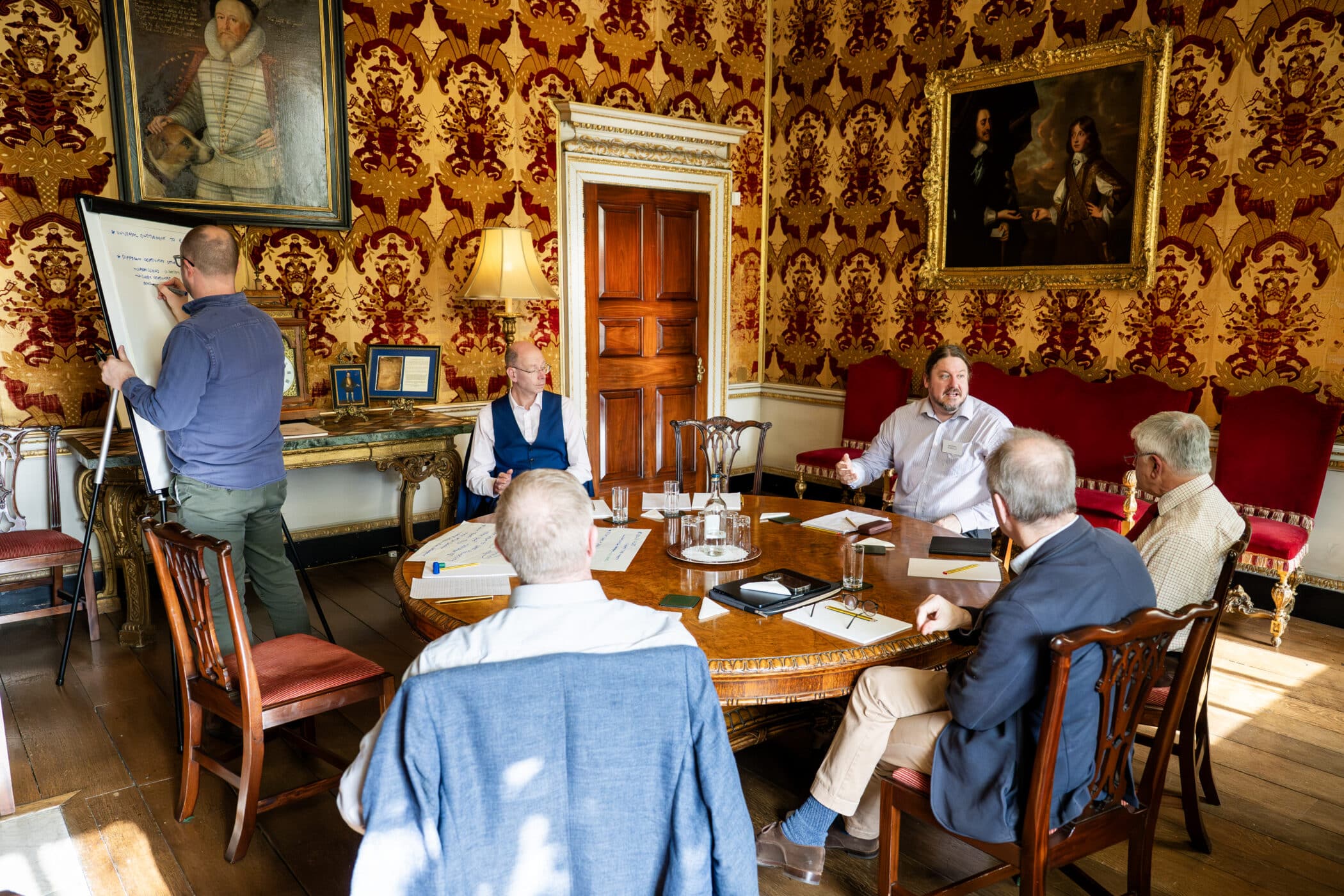 Six people are engaged in a meeting around a wooden table in an ornate room with red and gold wallpaper. One person stands writing on a flipchart, while others sit listening or discussing. The room features classical portraits, antique furniture, and warm lighting.