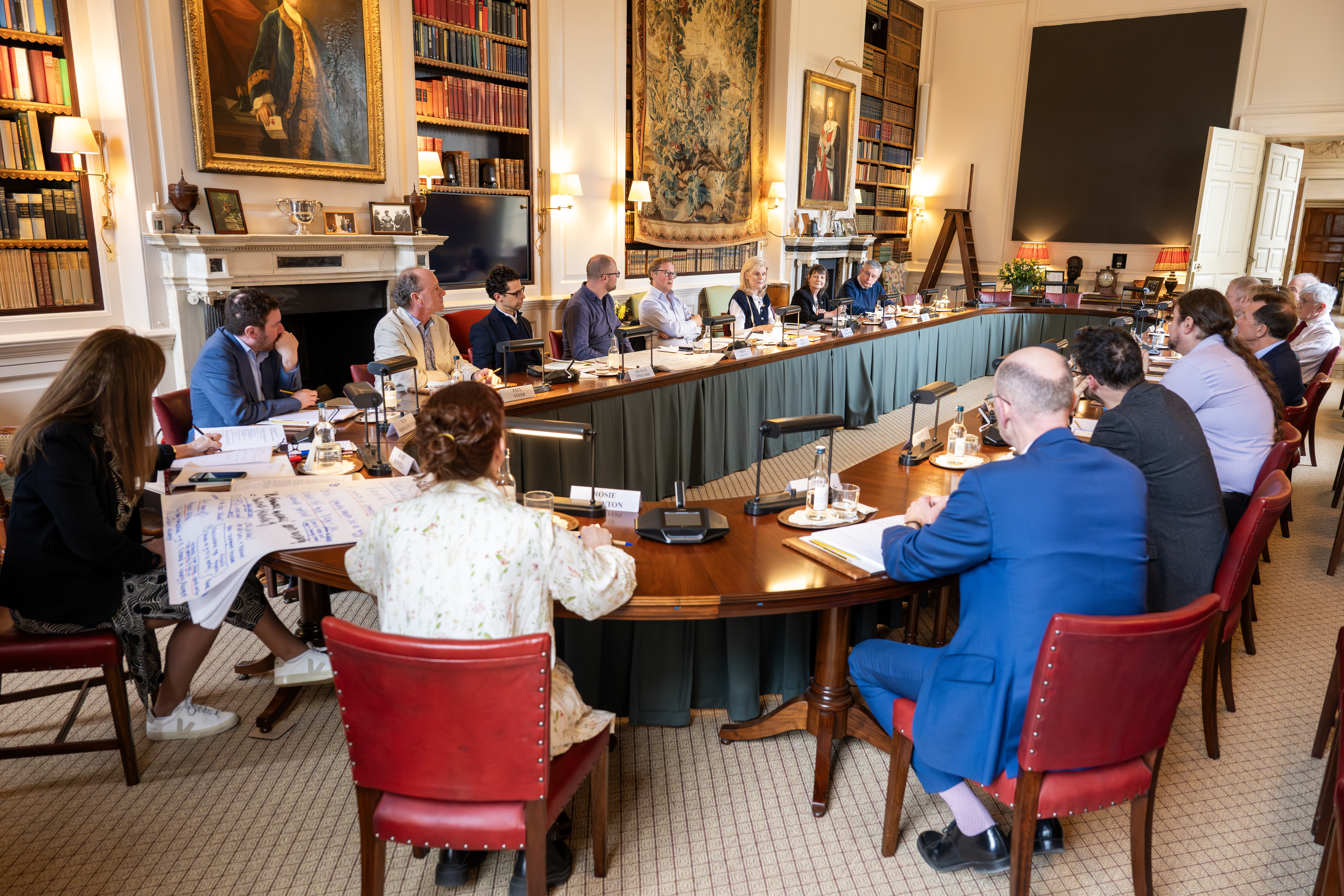 A large group of people sit around a U-shaped conference table in an ornate library-style room, engaged in discussion. The setting features red chairs, nameplates, notepads, and microphones, with bookshelves, portraits, and classical decor in the background.