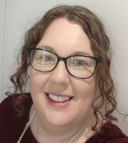A woman with fair skin, curly light brown hair, and black-framed glasses is smiling at the camera. She is wearing a burgundy top and a light-colored necklace. The background is a plain white wall.