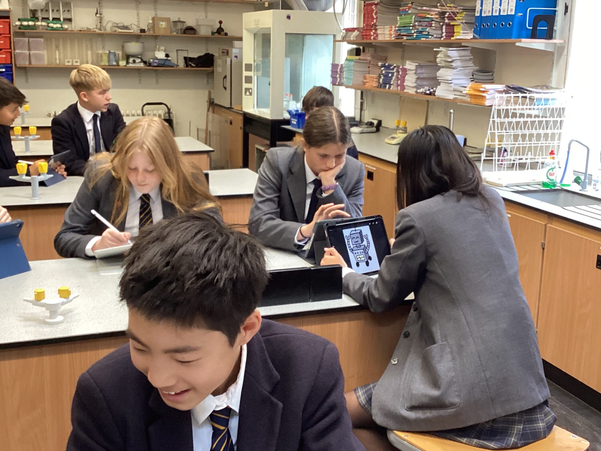 A group of students in school uniforms, consisting of blazers and ties, are engaged in a classroom activity in a science laboratory. Some students are writing on tablets with styluses, while others are discussing and collaborating. One student is holding a tablet displaying a digital drawing of a robot. The background includes lab equipment, shelves filled with books, and other educational materials, creating a dynamic and interactive learning environment.