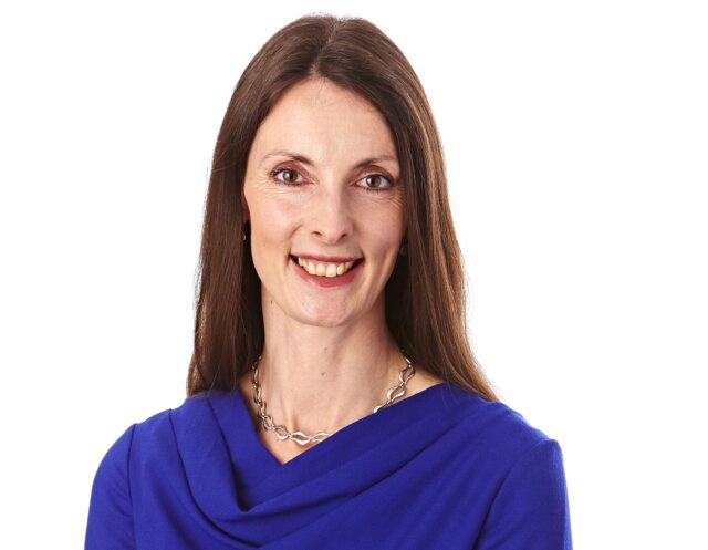 Woman with long brown hair smiling at the camera, wearing a royal blue dress with a draped neckline and a silver chain necklace, posed against a white background.