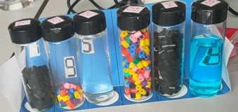 Six small transparent containers with black lids are arranged in a tray. Each container has a number or colorful contents inside, including black and multicolored beads, liquid solutions, and small objects.