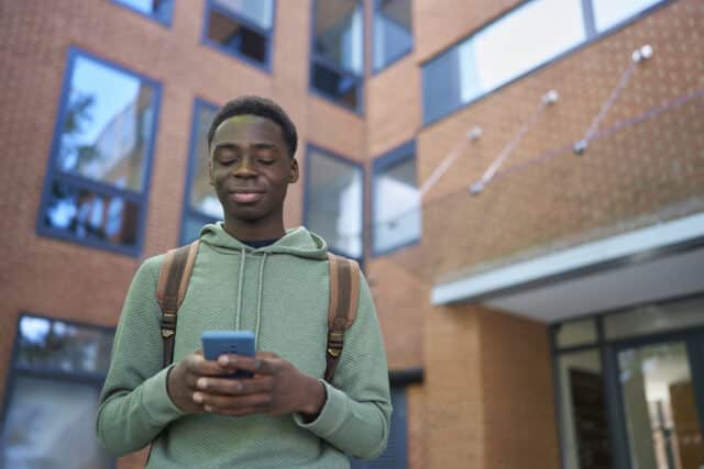 A young man wearing a green hoodie and a backpack stands outside a modern brick building with large windows. He holds a smartphone in both hands and has a calm, content expression with his eyes closed and a slight smile on his face. The background is slightly blurred, emphasizing the subject.