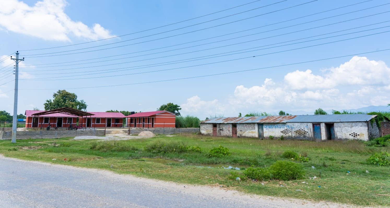 A photo of two school buildings in a rural setting in Nepal. On the left is a new red school building with a red roof, columns, and a shaded entry. On the right is an old, weathered, white stone building with a rusted metal roof. Both structures are surrounded by grassy fields under a blue sky with light clouds.