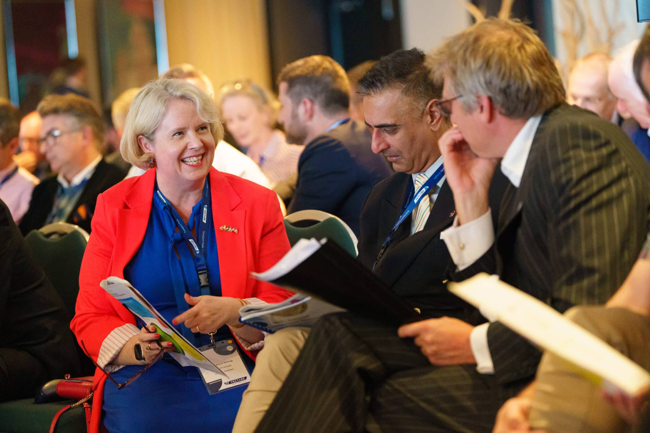 A group of professional delegates engaged in discussion during an HMC conference. A smiling woman in a red blazer and blue dress talks with two suited men, all wearing conference lanyards. They are seated among other attendees in a well-lit conference room, holding event materials.