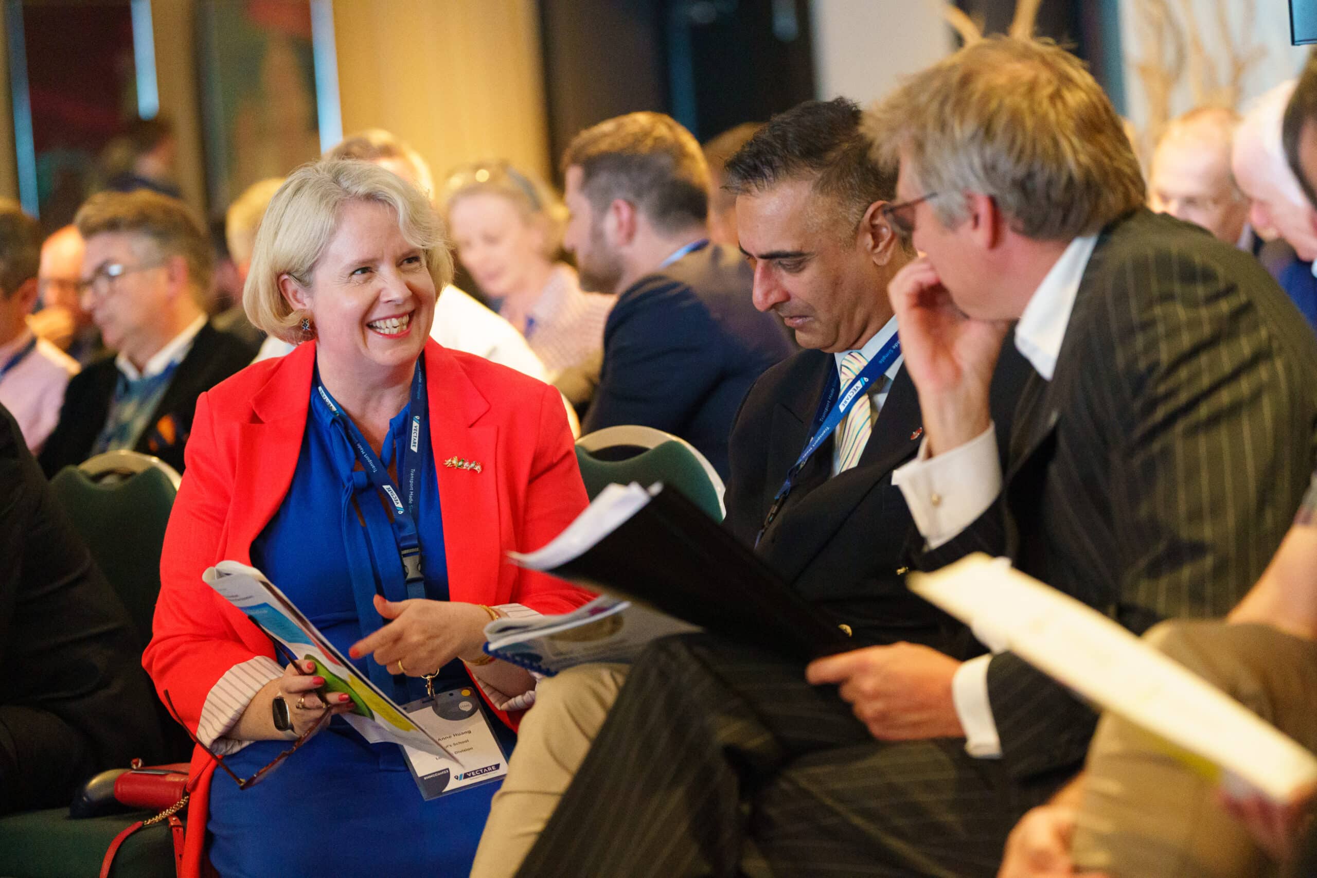 A group of professional delegates engaged in discussion during an HMC conference. A smiling woman in a red blazer and blue dress talks with two suited men, all wearing conference lanyards. They are seated among other attendees in a well-lit conference room, holding event materials.