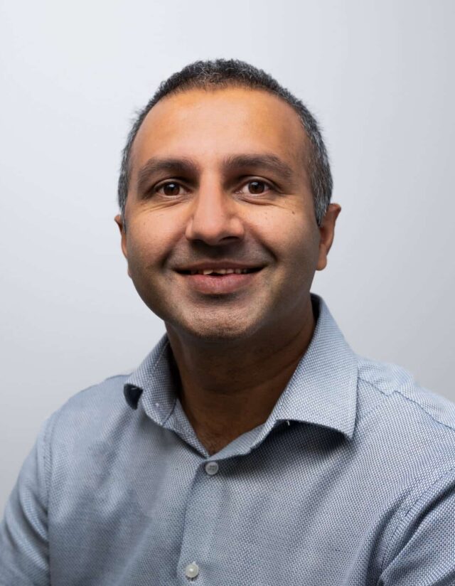 A smiling man with short, dark hair, wearing a light blue patterned shirt with an open collar. He faces the camera against a plain light grey background.