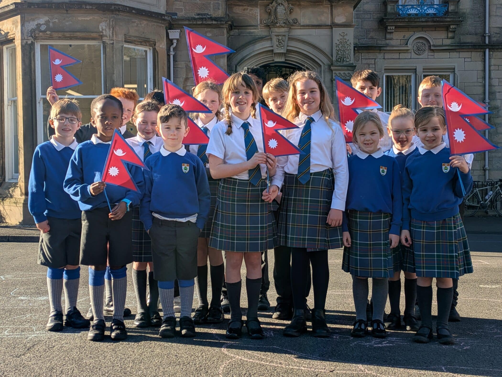 A group of schoolchildren in blue sweaters and plaid uniforms stand outside a stone school building, smiling and holding Nepalese flags. The children are standing in sunlight, and the setting appears to be a school courtyard.