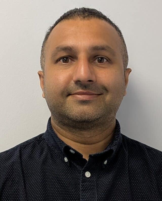 A man with short dark hair and a trimmed beard is facing the camera and smiling slightly. He is wearing a dark blue button-up shirt with a subtle dotted pattern. The background is a plain white wall.