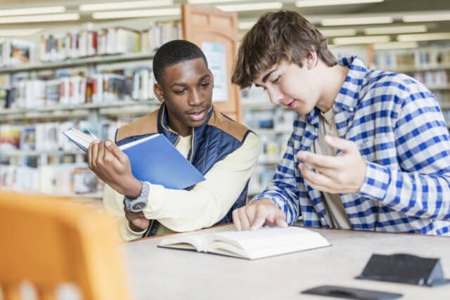 Two teenage students studying together at a library table. One holds a blue book while the other gestures toward an open textbook on the table. Bookshelves filled with books are visible in the background.
