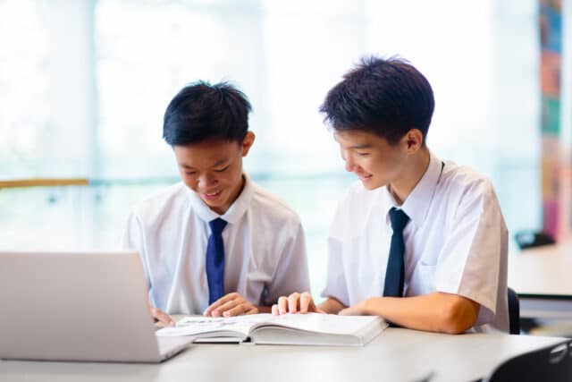 Two male students in school uniforms, smiling and working together at a desk with an open book and a laptop. They are seated in a bright, modern classroom or library setting.