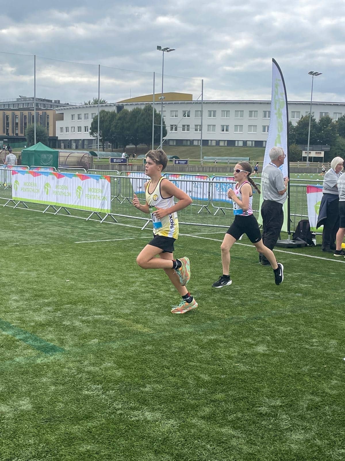 Young athletes running on green turf, wearing race bibs and yellow/black uniforms. Spectators and officials stand by.