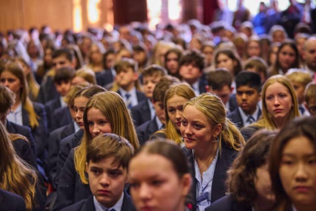 A large group of secondary school students dressed in formal uniforms are seated closely together, attending an assembly in what appears to be a school hall. Most students are focused ahead, with some showing attentive or thoughtful expressions. The lighting is warm, and the depth of the image emphasizes the size of the gathering, with rows of students fading into the background.