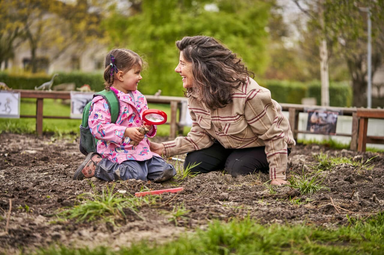 A young girl and an adult woman kneeling in a muddy outdoor area, smiling at each other. The girl is holding a red magnifying glass and a toy dinosaur, wearing a pink raincoat with owl prints and a green backpack. Behind them are printed dinosaur images attached to a wooden frame, suggesting an educational or play activity related to paleontology.