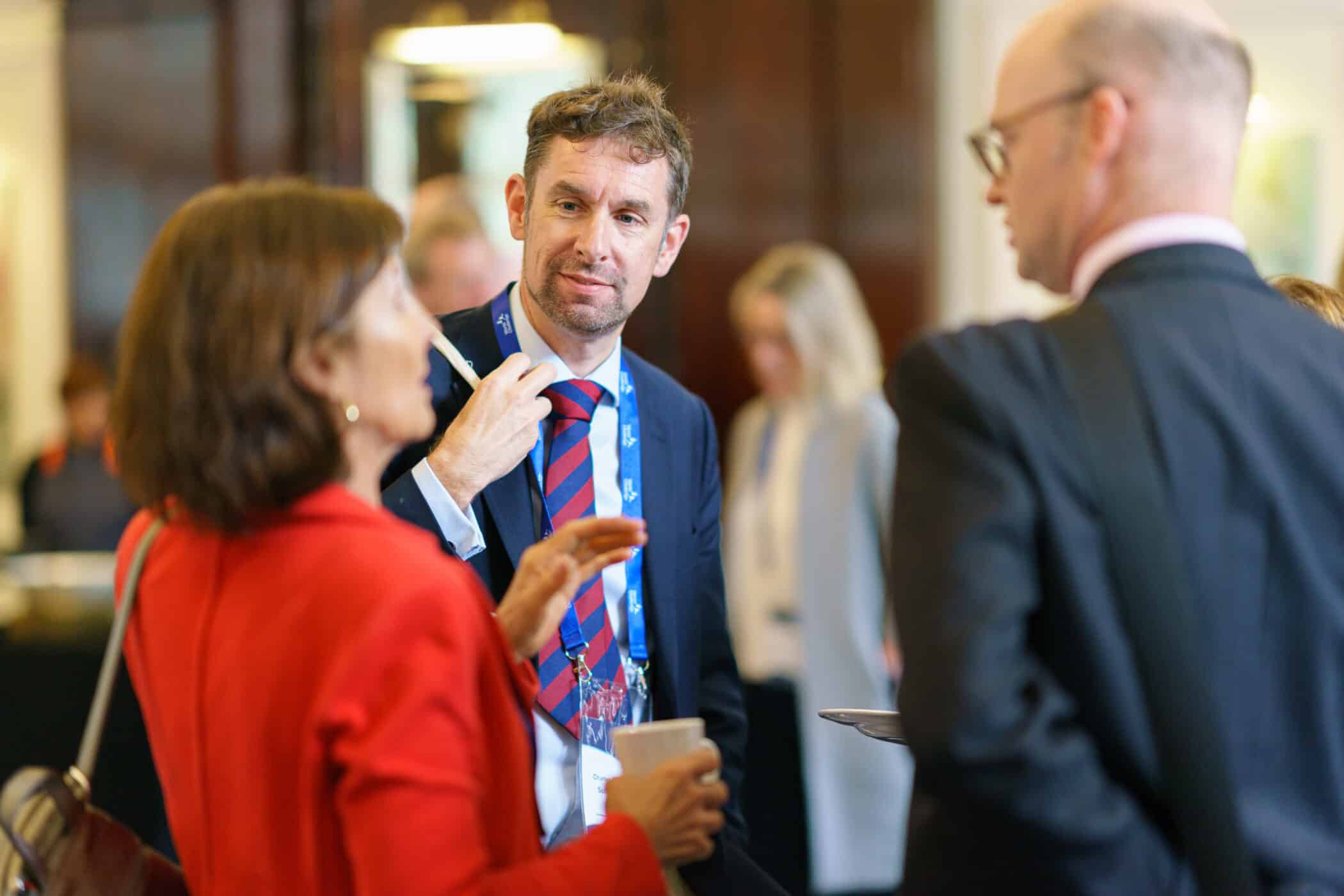 Three professionally dressed individuals engage in conversation at what appears to be a formal networking or conference event. The man in the center, wearing a navy suit, red and blue striped tie, and a blue lanyard, is attentively listening to the woman in red on the left, who is speaking and holding a coffee cup. Another man, bald and wearing glasses, is partially visible on the right. The background is softly blurred, showing other attendees and a polished interior, suggesting a conference or business gathering.
