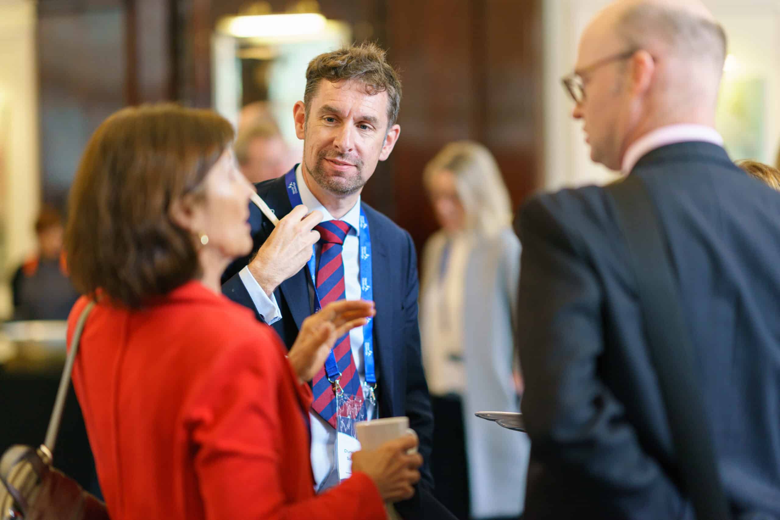 Three professionally dressed individuals engage in conversation at what appears to be a formal networking or conference event. The man in the center, wearing a navy suit, red and blue striped tie, and a blue lanyard, is attentively listening to the woman in red on the left, who is speaking and holding a coffee cup. Another man, bald and wearing glasses, is partially visible on the right. The background is softly blurred, showing other attendees and a polished interior, suggesting a conference or business gathering.