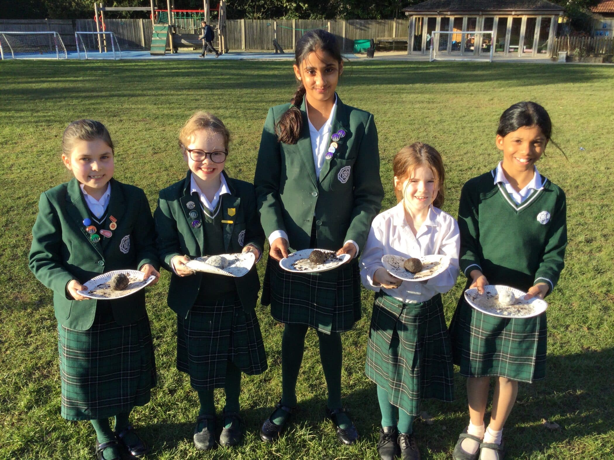 A group of students in school uniforms standing in a grassy outdoor area, holding paper plates with mud balls or seed bombs, participating in an eco-friendly learning activity.