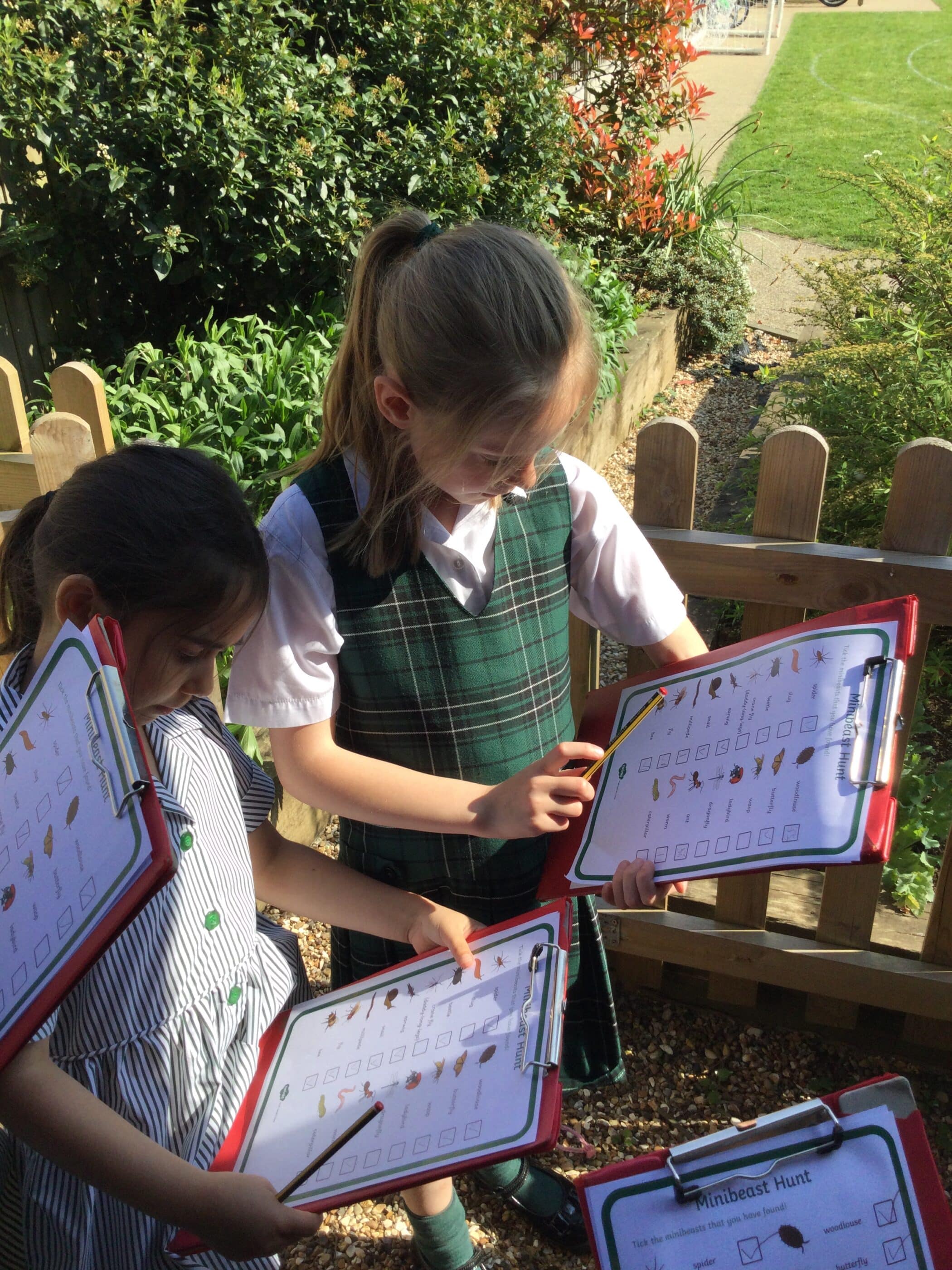 Young students sitting on benches outside, holding clipboards and papers, actively engaged in a field study or survey task. They are smiling and focused, wearing green school uniforms.