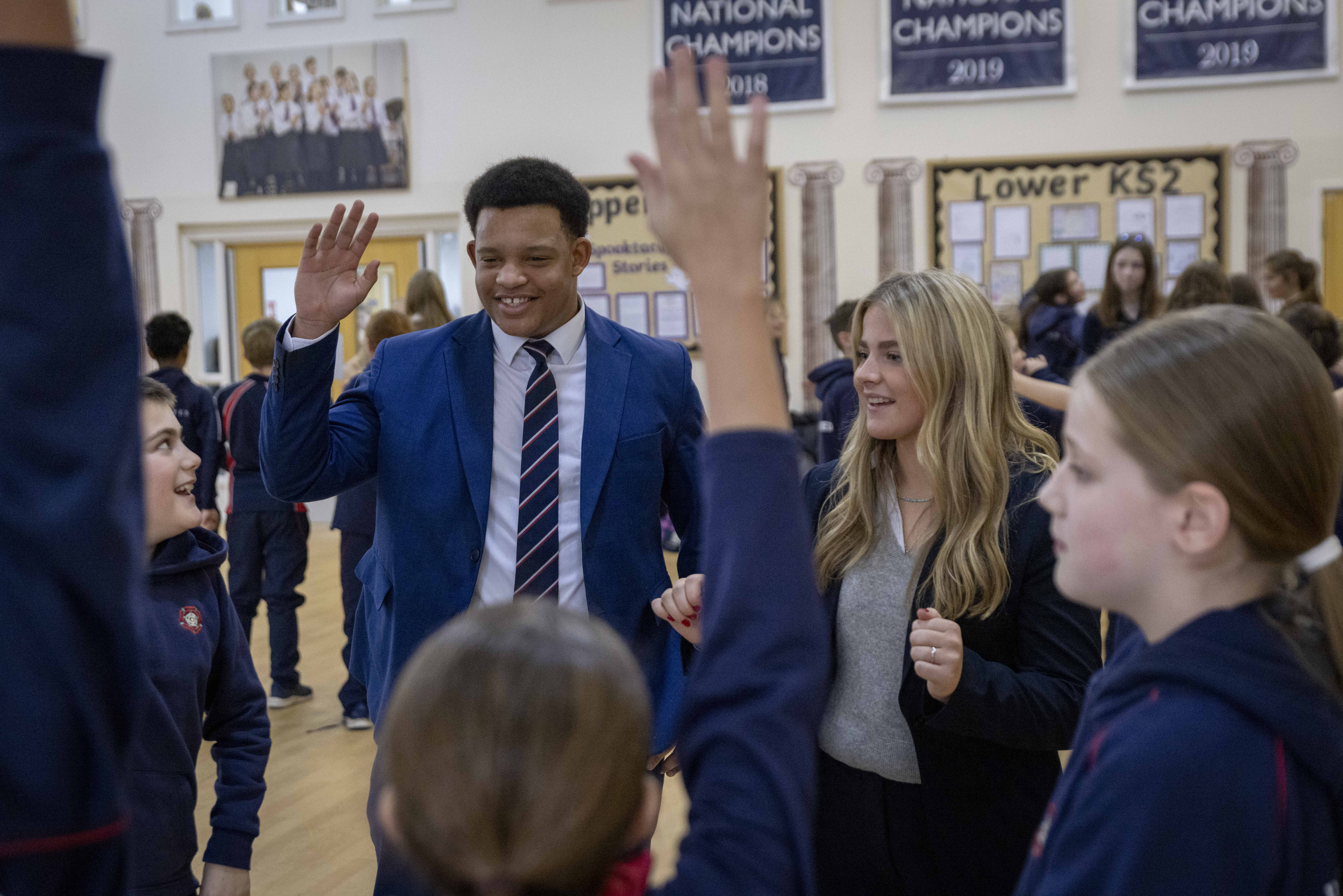 A male and female sixth form student in formal dress stand in the middle of a school hall interacting with a group of younger pupils. The male student smiles broadly and raises his hand, as several children raise theirs enthusiastically. Display boards and banners showing “National Champions” hang in the background.