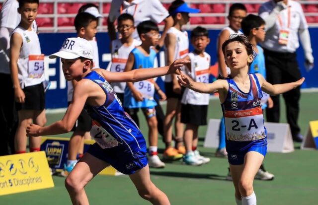 Two boys in blue uniforms pass a baton in a relay race, while other kids in race bibs watch.