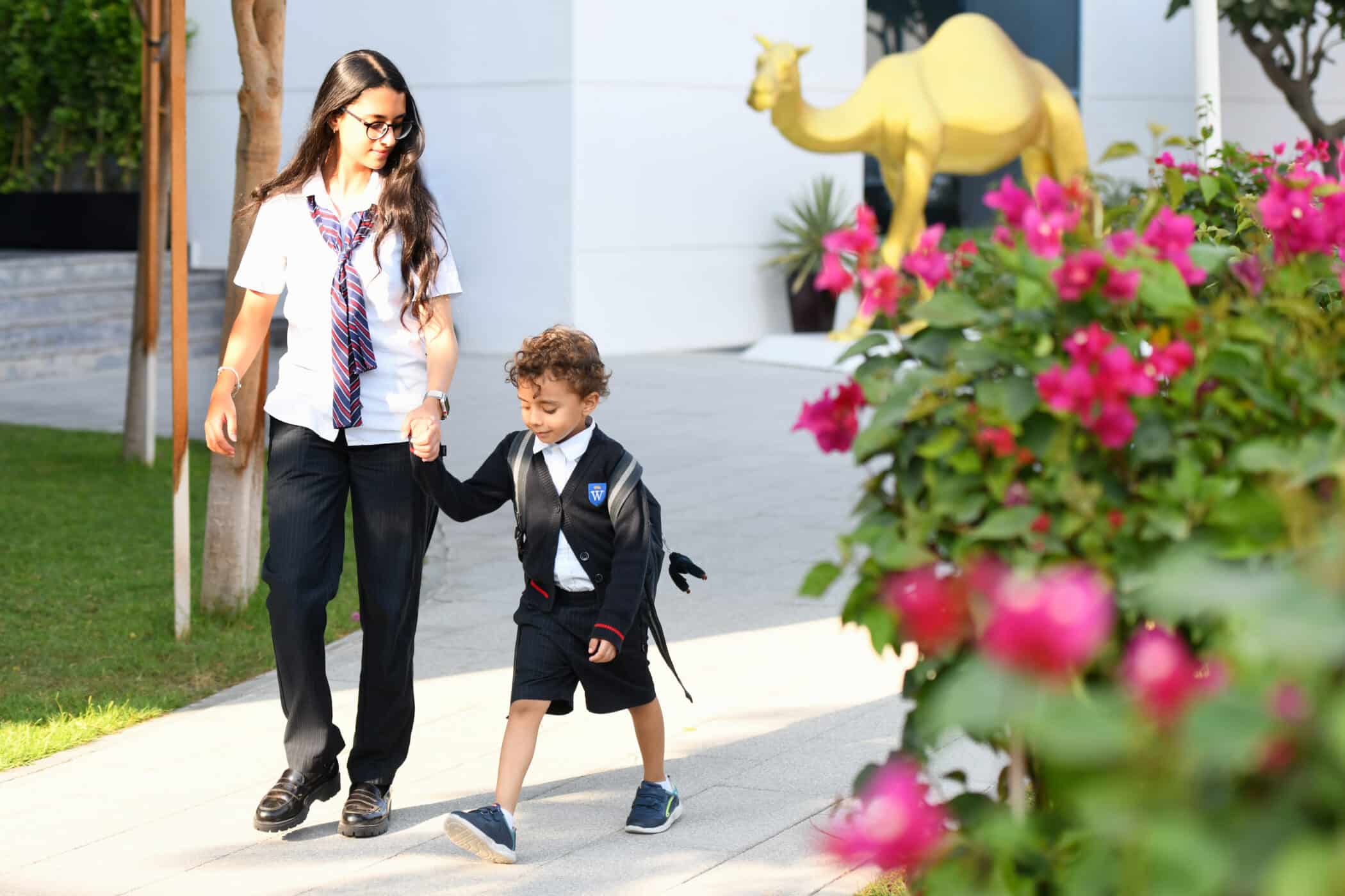 A young woman in a school uniform, wearing glasses and a striped tie, walks hand-in-hand with a small boy in a matching school uniform with a crest on his sweater. They are walking on a paved path beside a green lawn and a flowering bush with pink blossoms. In the background, there is a golden camel statue and a modern white building. Both appear cheerful and focused on their walk.
