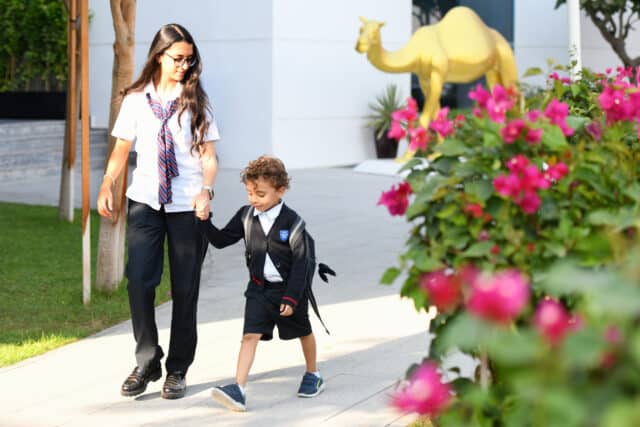 A young woman in a school uniform, wearing glasses and a striped tie, walks hand-in-hand with a small boy in a matching school uniform with a crest on his sweater. They are walking on a paved path beside a green lawn and a flowering bush with pink blossoms. In the background, there is a golden camel statue and a modern white building. Both appear cheerful and focused on their walk.