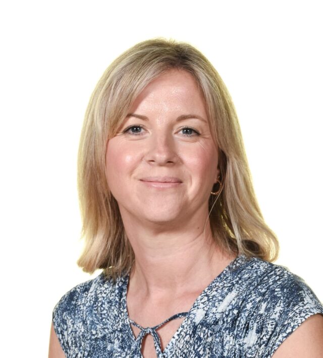 Professional portrait of a smiling woman with shoulder-length blonde hair, wearing a blue patterned blouse. She is posed against a plain white background.