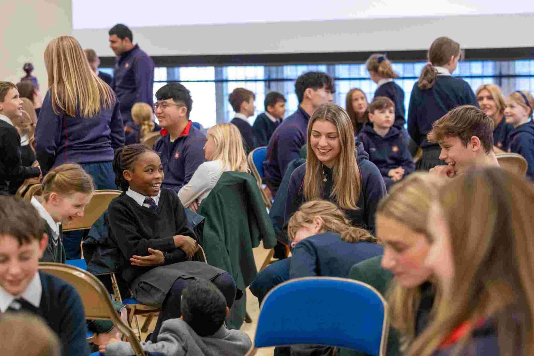 A large group of students from different year groups interact in a lively school hall. Most of the older students wear navy hoodies with red trim, while younger students are in uniform. Small groups are seated in folding chairs, engaged in warm conversation, with smiles and laughter visible.