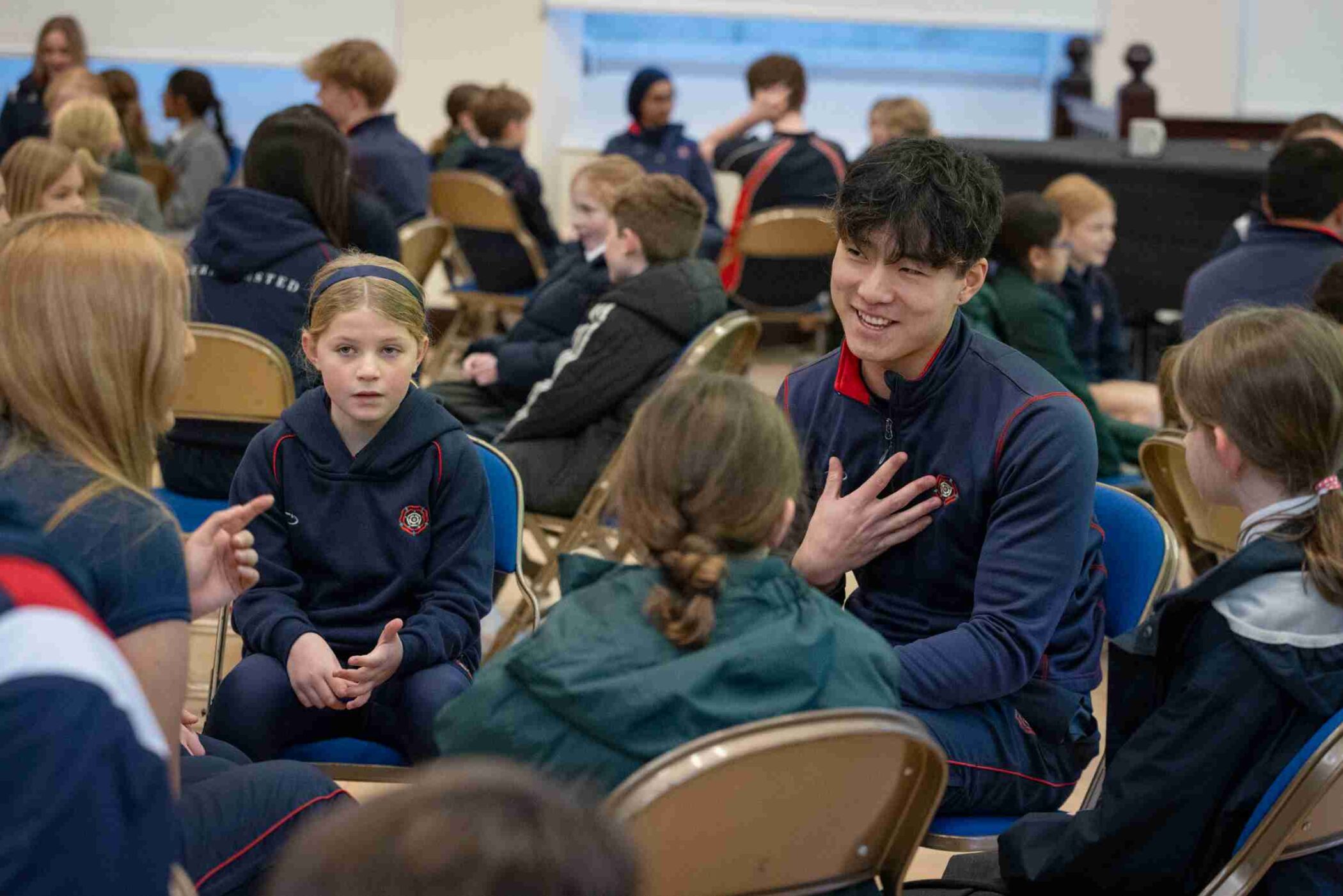 Students of various ages sit in small discussion circles on folding chairs in a school hall. A boy in a navy tracksuit with red trim smiles as he speaks to younger pupils. A girl in a headband sits nearby listening attentively, while others engage in the background.
