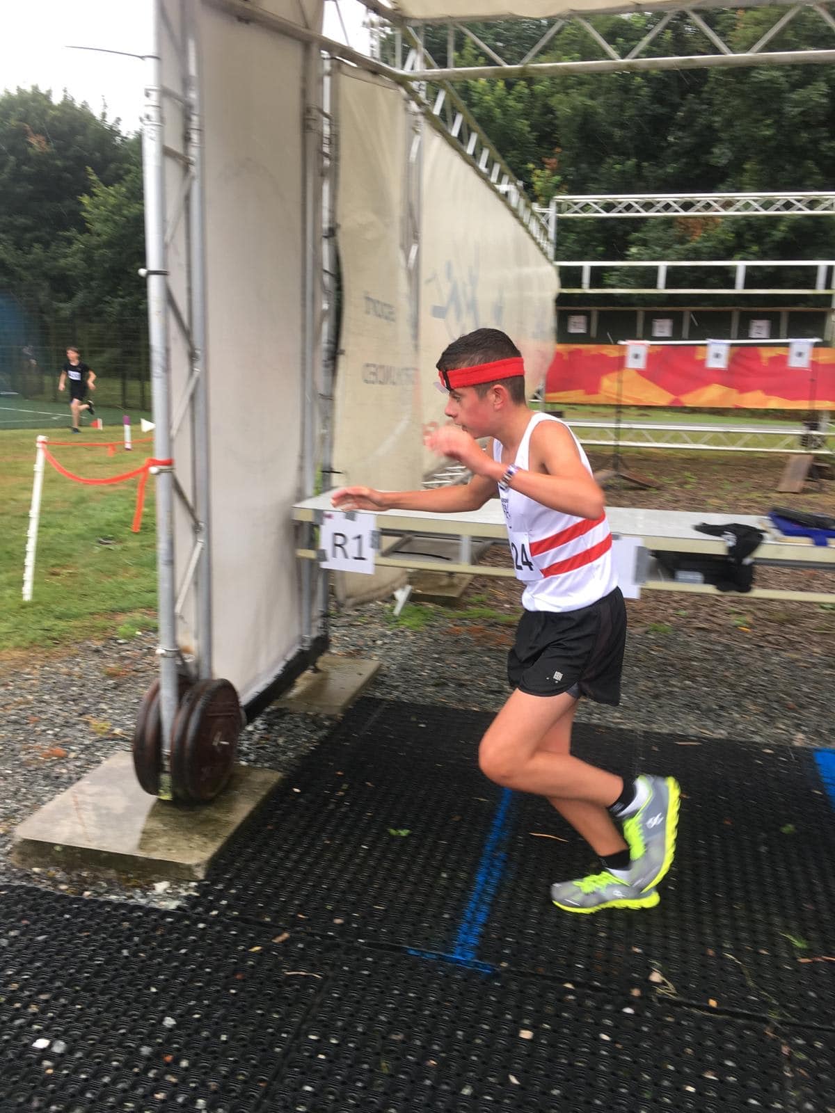 Teenage boy in a red headband and white/red outfit runs through a gated area in a pentathlon event, with targets in the background.