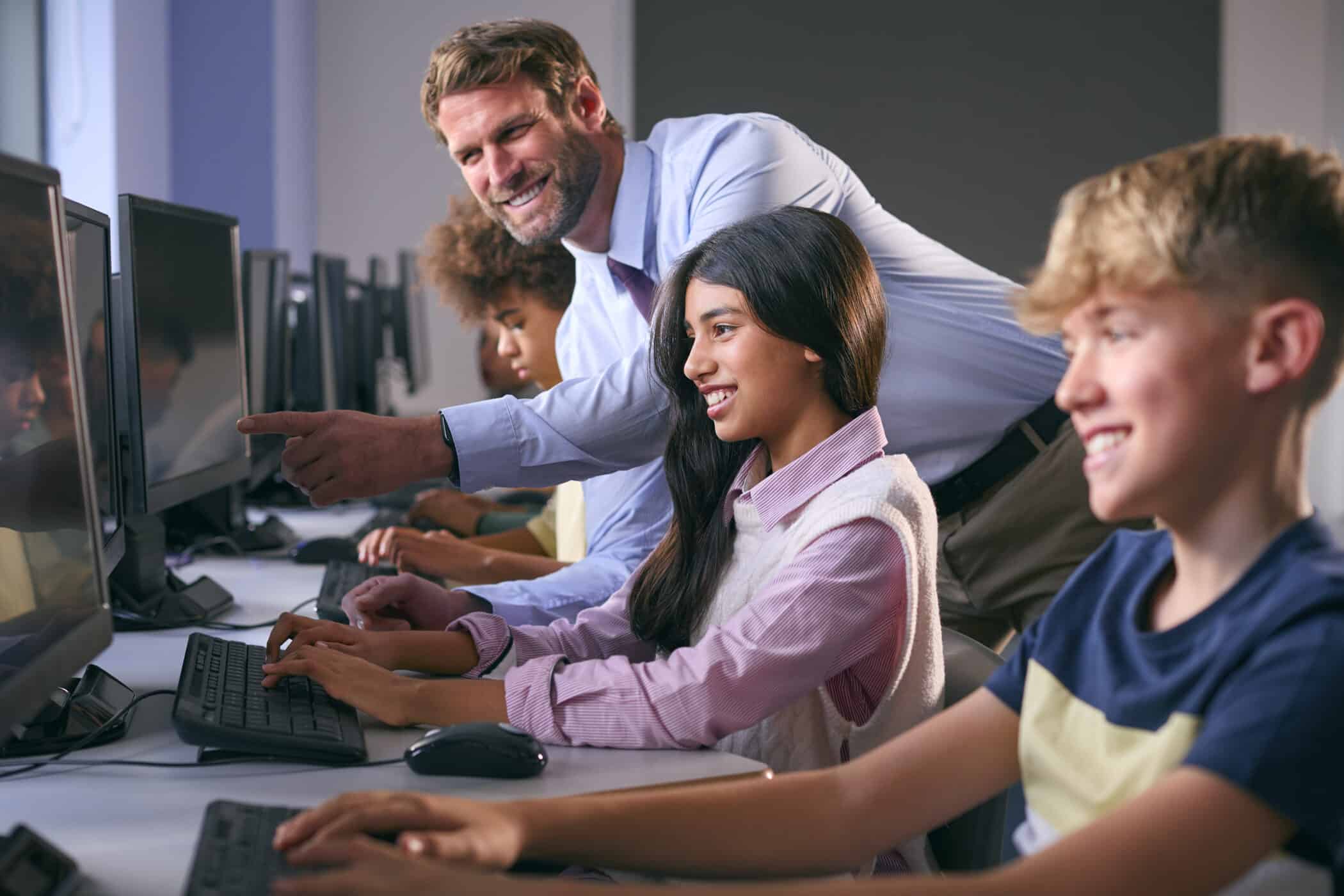 A series of images depicting a computer science classroom environment. A male teacher with short brown hair, wearing a light blue shirt and tie, is actively engaging with students seated at desktop computers. The students, a diverse group of boys and girls in their early teens, are focused on their computer screens, smiling, typing, and working together in a collaborative learning setting. The classroom has rows of computers and a bright, modern feel.