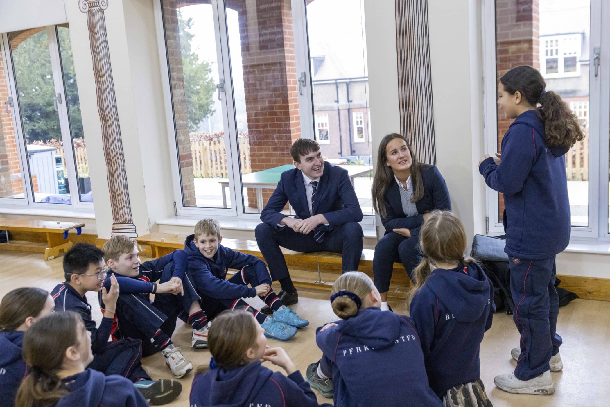 A group of schoolchildren in navy tracksuits sit on a wooden floor in a bright, spacious room with tall windows. Two sixth form students in formal attire—navy suits and ties—engage attentively with the group, while a young girl stands speaking to them. Everyone appears involved and cheerful.