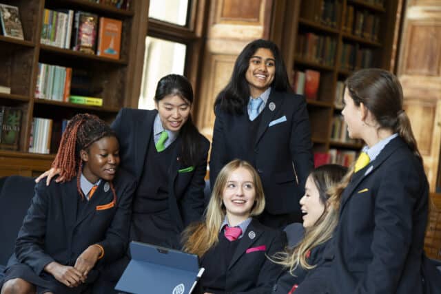 A diverse group of six female students in school uniforms, gathered around a tablet and smiling. Each student is wearing a blazer with a striped shirt and a different coloured tie and pocket square, suggesting house or group distinctions. They are seated and standing in a wood-panelled library with shelves of books in the background.