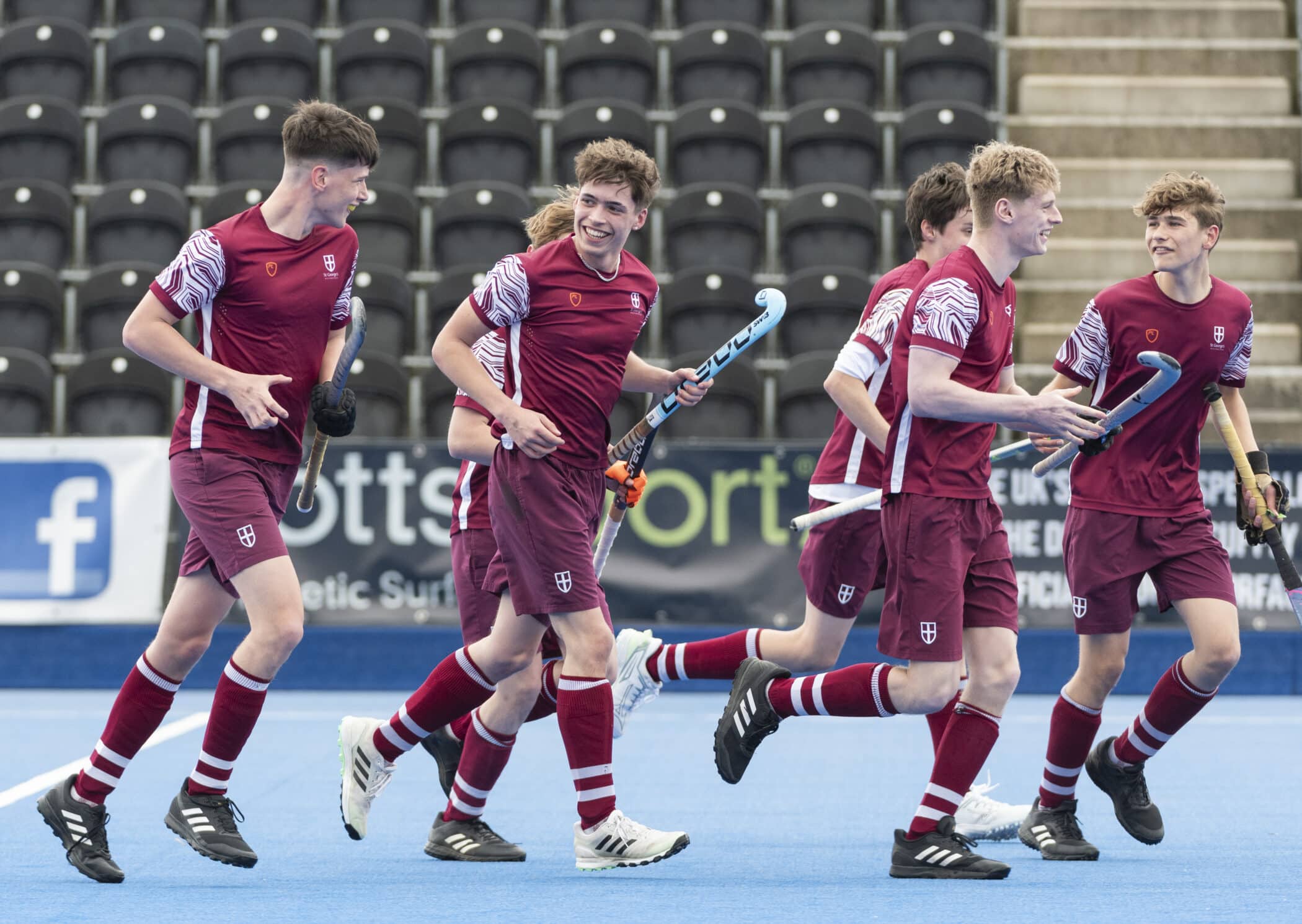 A group of teenage boys in coordinated maroon hockey uniforms run together smiling and laughing on a blue artificial pitch, holding hockey sticks, likely after scoring a goal during a match.
