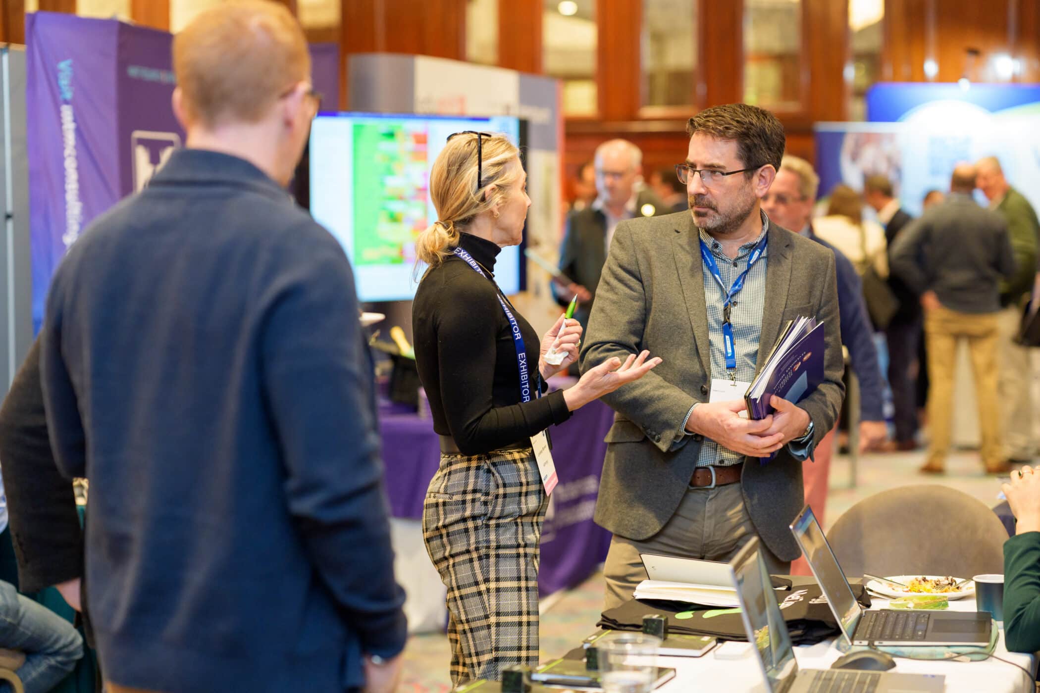 People networking at a conference exhibition area, with attendees and exhibitors engaged in conversation beside display booths and tables with laptops and promotional materials.